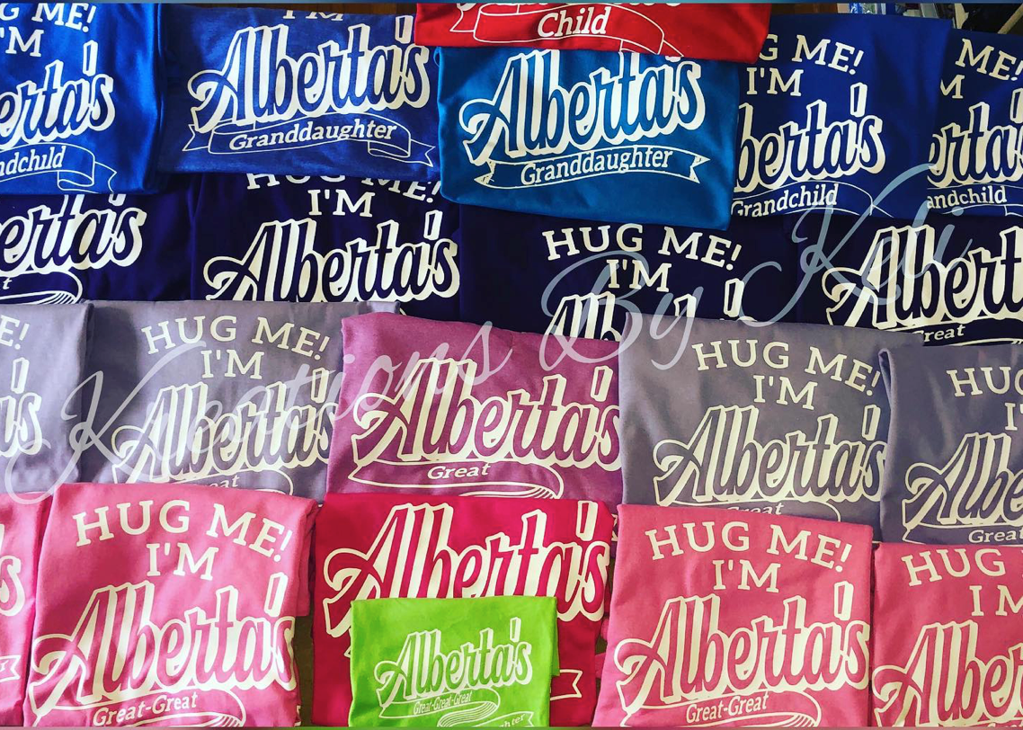 A collection of family reunion t-shirts in various colors, each with a name printed on the front and the phrase 'HUG ME'.